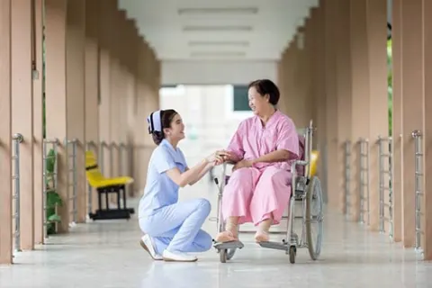Nurse crouching to speak with a patient seated in a wheelchair in a hospital corridor